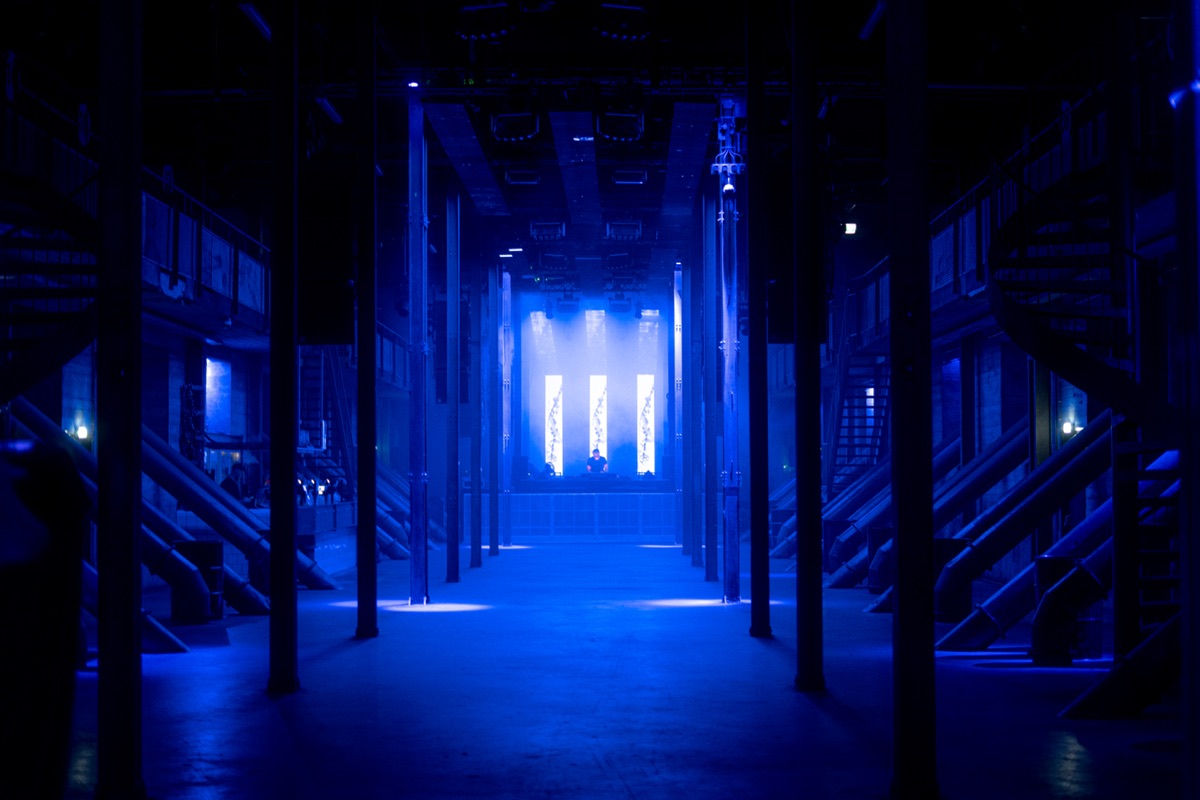 a symmetrical concrete hall in deep blue light. three led panels hang from the ceiling and six more line the back wall in two rows, a dj lit small between them at the far end.